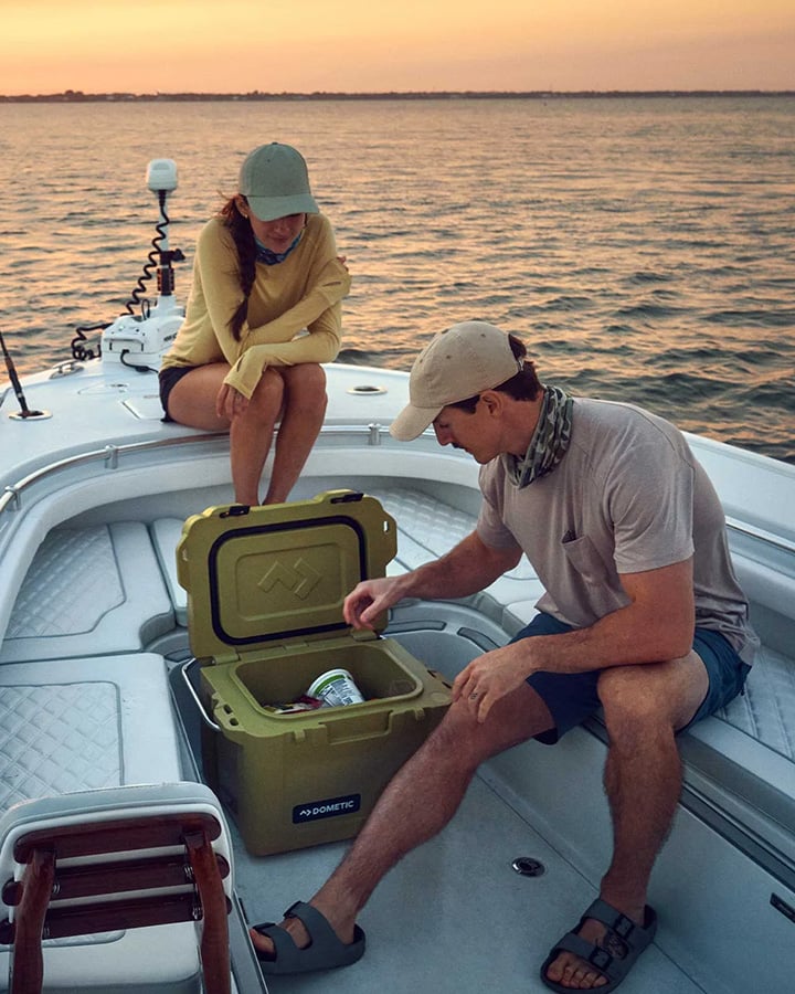 Dwie osoby na łodzi o zachodzie słońca z otwartą zieloną lodówką Dometic na pokładzie, w której znajdują się napoje.