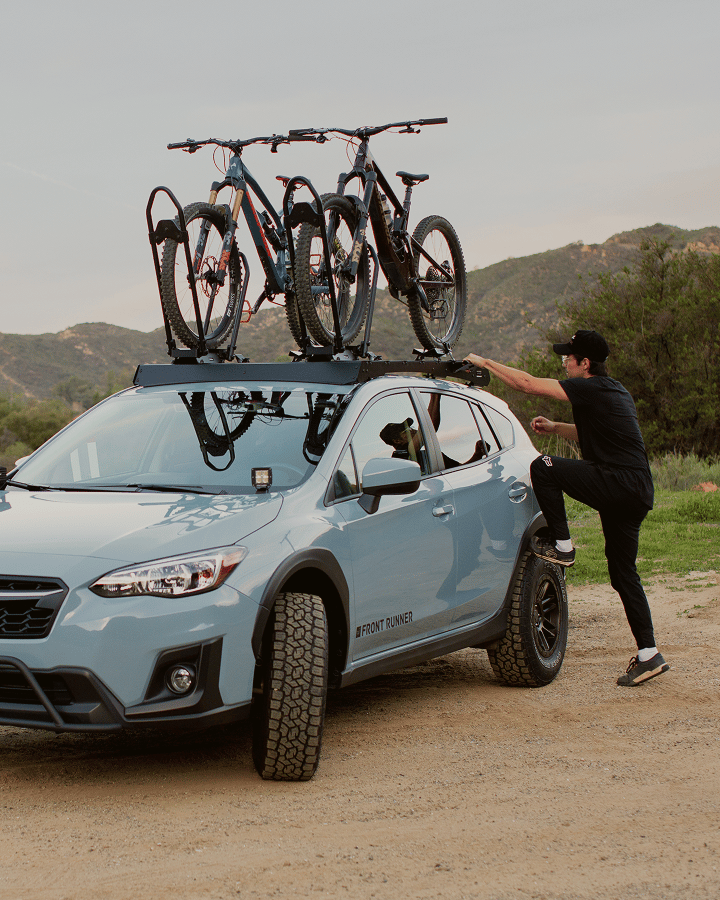 Véhicule avec des vélos montés sur une galerie de toit tandis qu’une personne sécurise les vélos en extérieur.