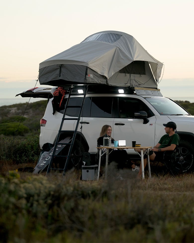 Véhicule blanc près de la côte au coucher du soleil avec une tente de toit Front Runner Dometic Feather‑Lite II installée, échelle fixée et table de camping à côté du véhicule.