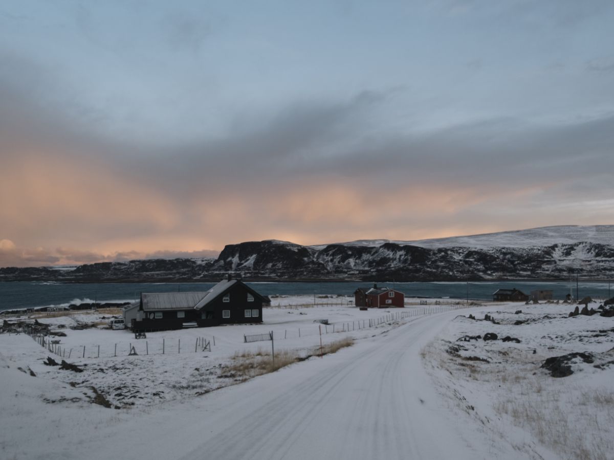 Snow covered road winding through a remote coastal landscape with scattered houses near the ocean and pink, orange sky.ear the