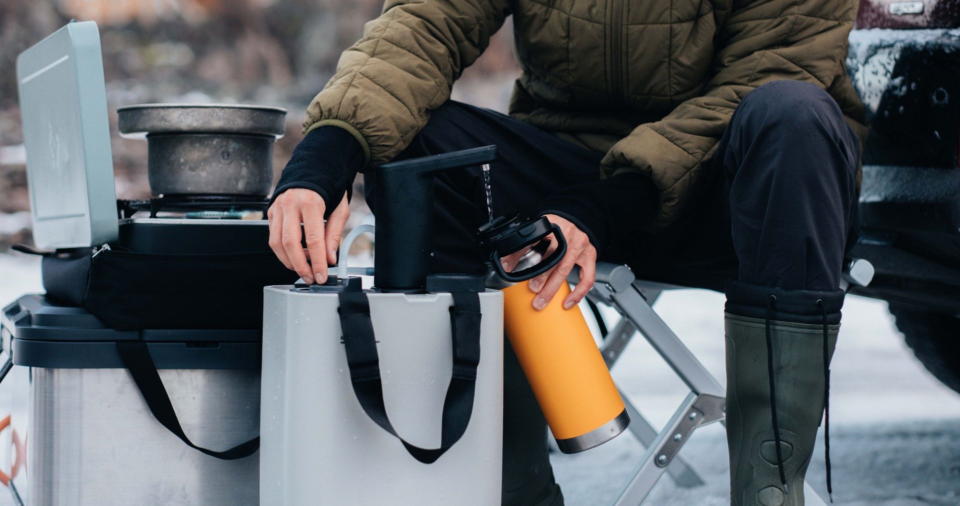 A person sitting down refilling a drinkbottle from the dometic hydration kit while a camping stove with a pot is set up nearby..