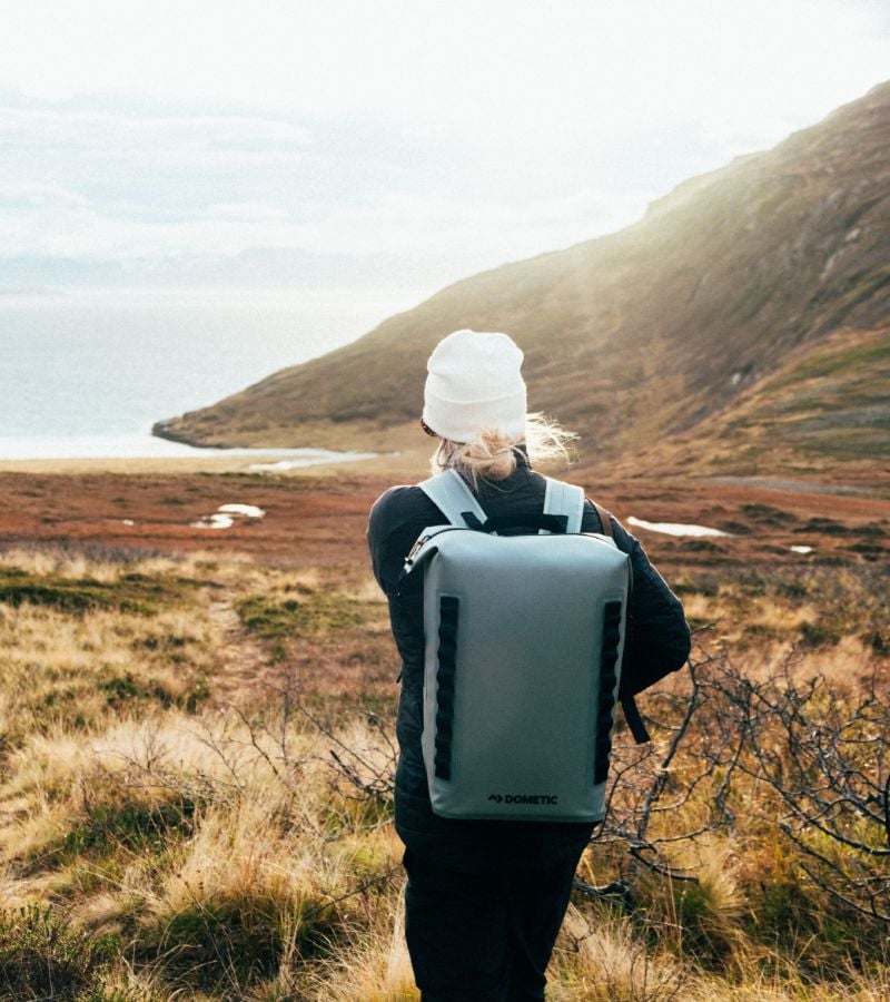 A female standing on a grassy hill overlooking a coastal landscape with a Dometic backpack.