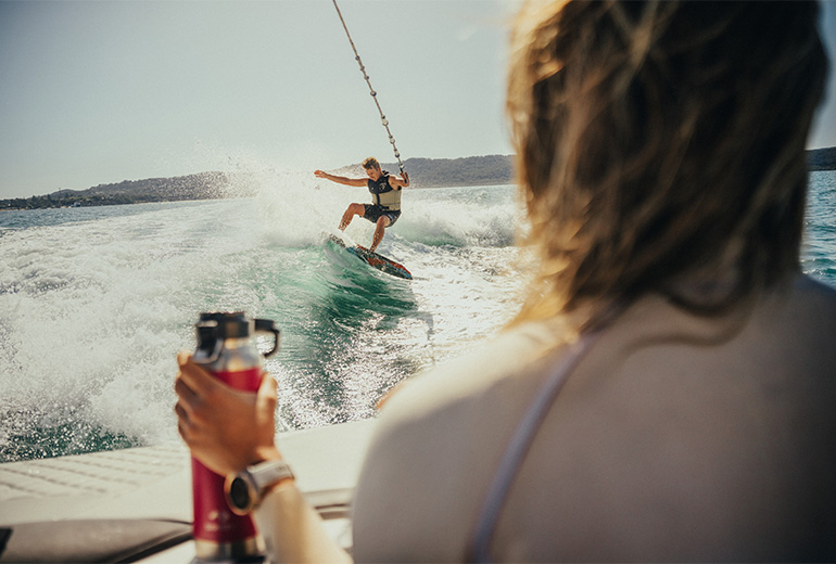 A woman holding a thermo bottle on a boat looking at a man that is wakeboarding 