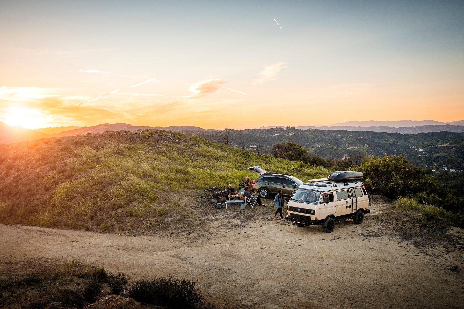 An old school yellow VW van and a dark green subaru parked at the top of a hill with a group of friends watching sunset and enjoying food an drinks at a Dometic table.