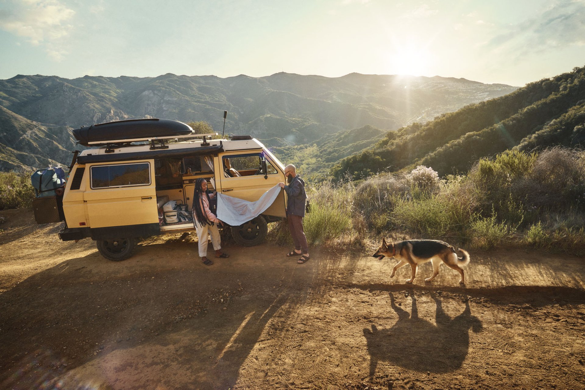 A man and woman folding a blanket outside their old school yellow VW camper van, with a german sherpard dog running near by.