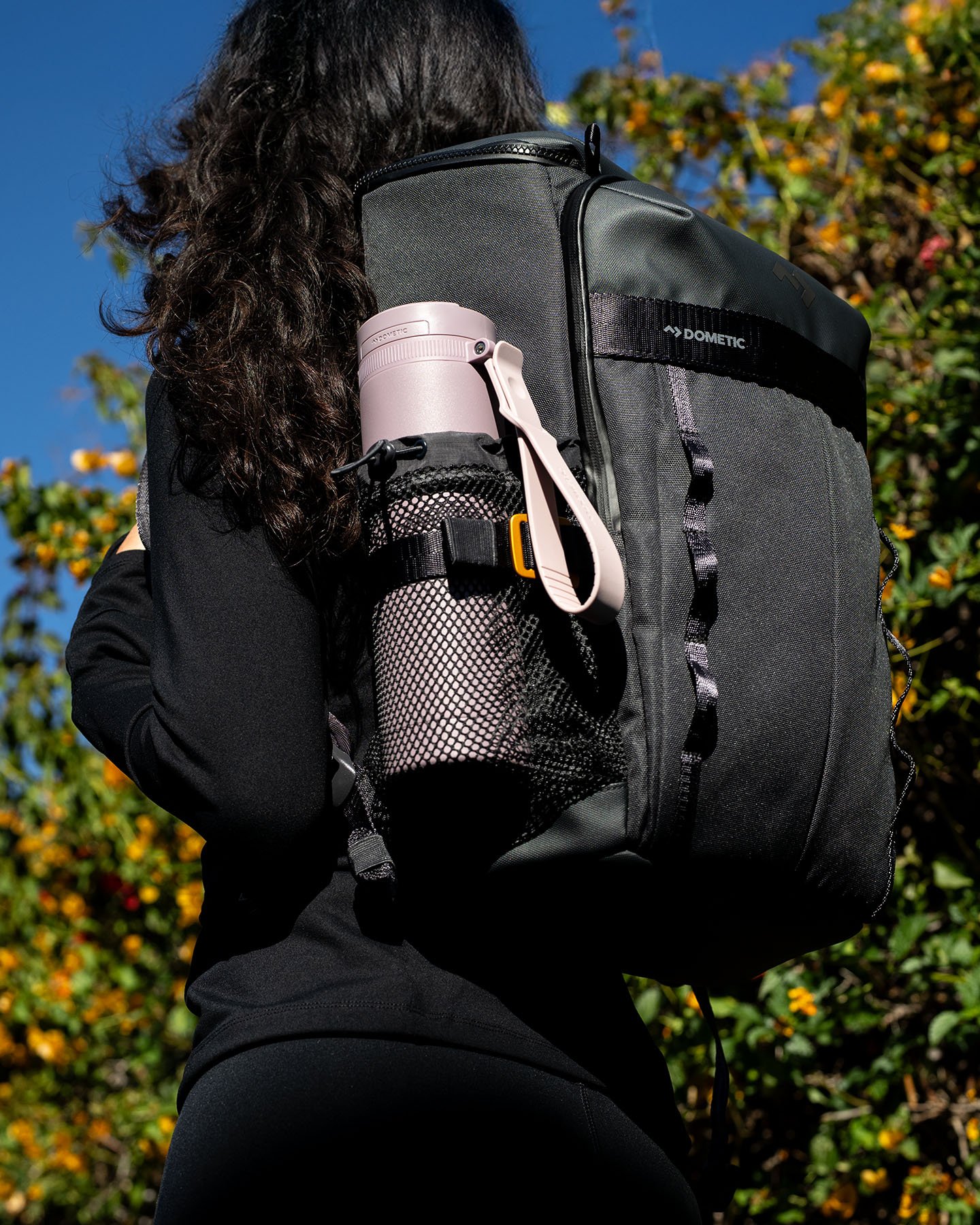 Persona con uno zaino Dometic nero all’aperto, con una borraccia isolata rosa nella tasca laterale in rete.