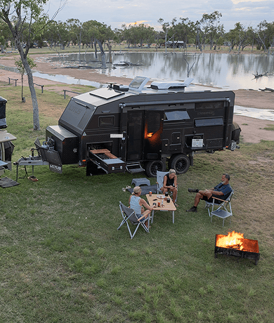 Off-road camper trailer with Dometic RV air conditioner parked beside a lake at dusk, with a campfire and folding chairs.