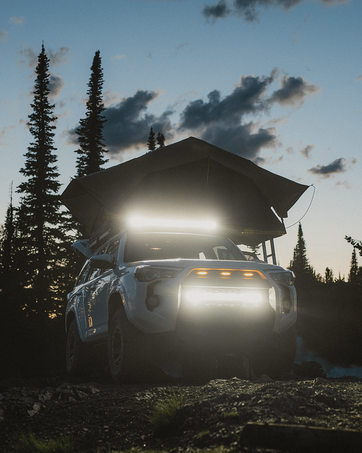 Off‑road SUV with a rooftop tent and bright LED lights parked in a forest at dusk.