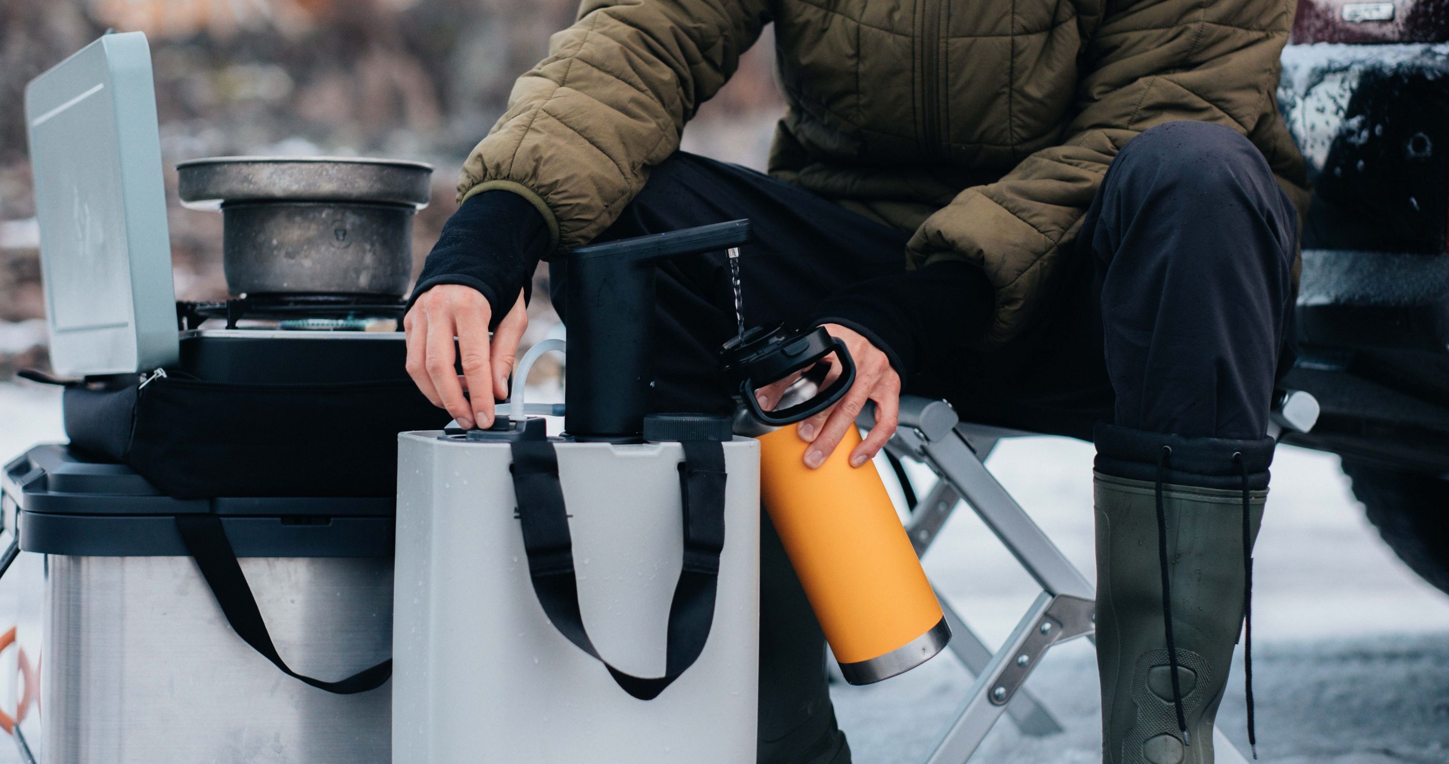 A person sitting down refilling a drinkbottle from the dometic hydration kit while a camping stove with a pot is set up nearby..