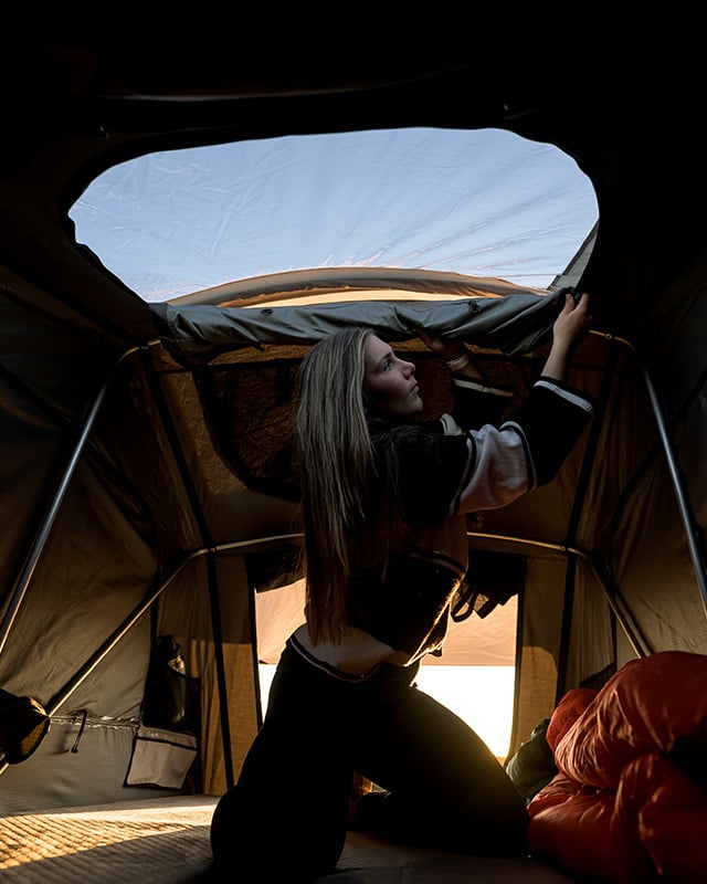 Person inside an opened Front Runner Dometic Feather‑Lite II rooftop tent adjusting the stargaze roof window, with a sleeping bag visible inside.
