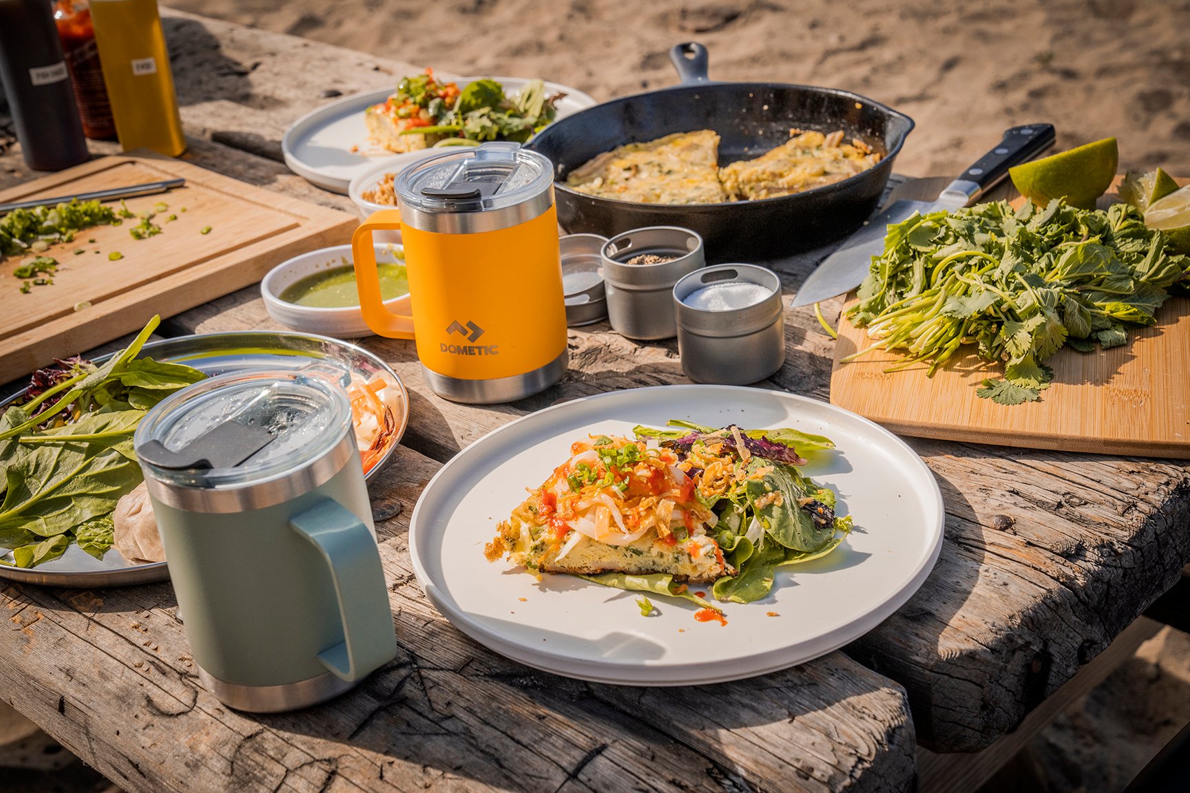 A close up of plates with prepared food on them and Dometic mugs on a picnic table.