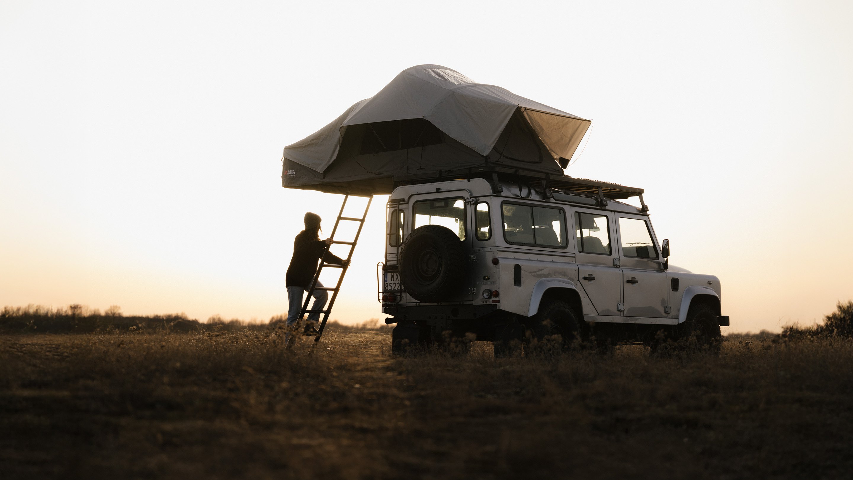 Véhicule tout-terrain avec tente de toit dans un paysage ouvert au coucher du soleil, personne montant à une échelle arrière pour accéder au camping surélevé