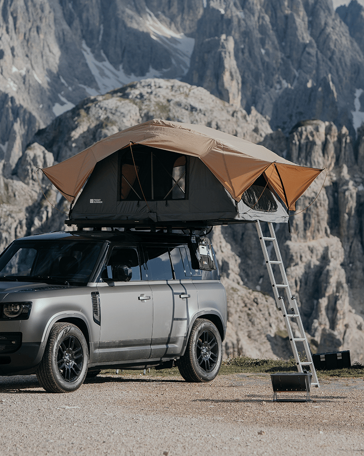 Pickup truck with a rooftop tent and ladder set up in a grassy outdoor area.
