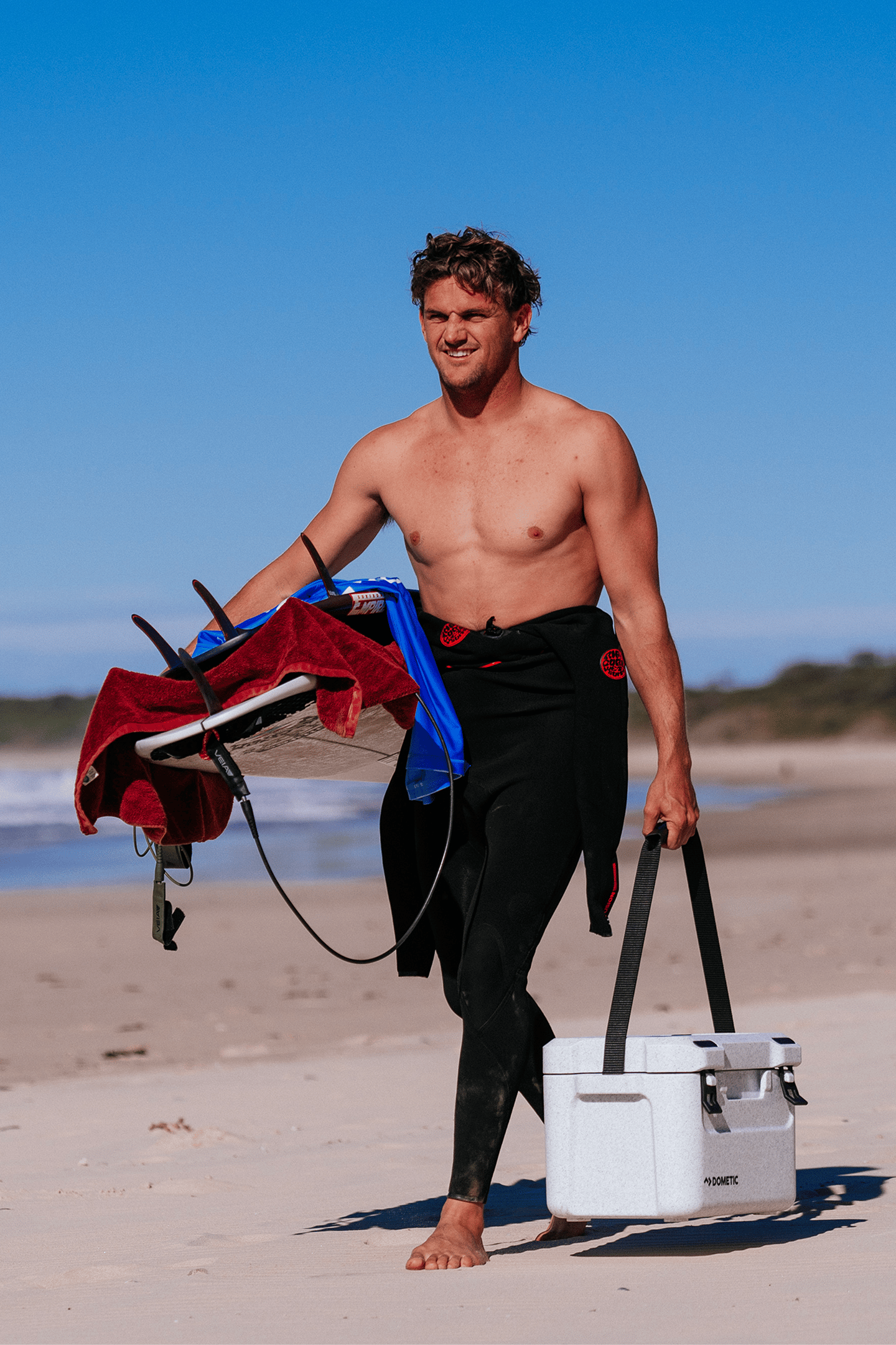 A surfer walks along the beach with a Dometic Cool-Ice CI 15 portable cooler in one hand and a surfboard in the other, ready for a day on the water. Compact, insulated and built to keep drinks and food cold for every coastal camping beach day