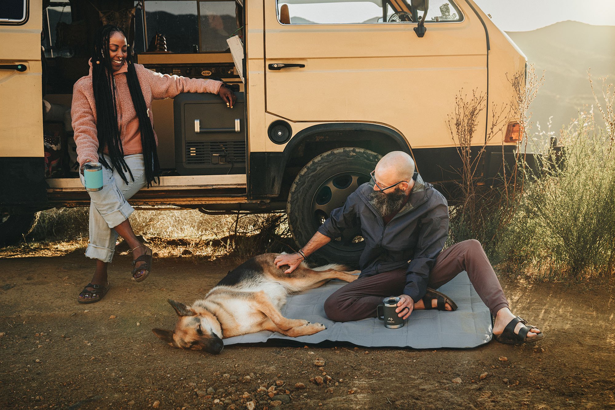 A woman sitting on the floor of an old school yellow VW van with a Dometic mug in her hand. A man sitting on a Dometic blanket with german shepard dog, next to the van.