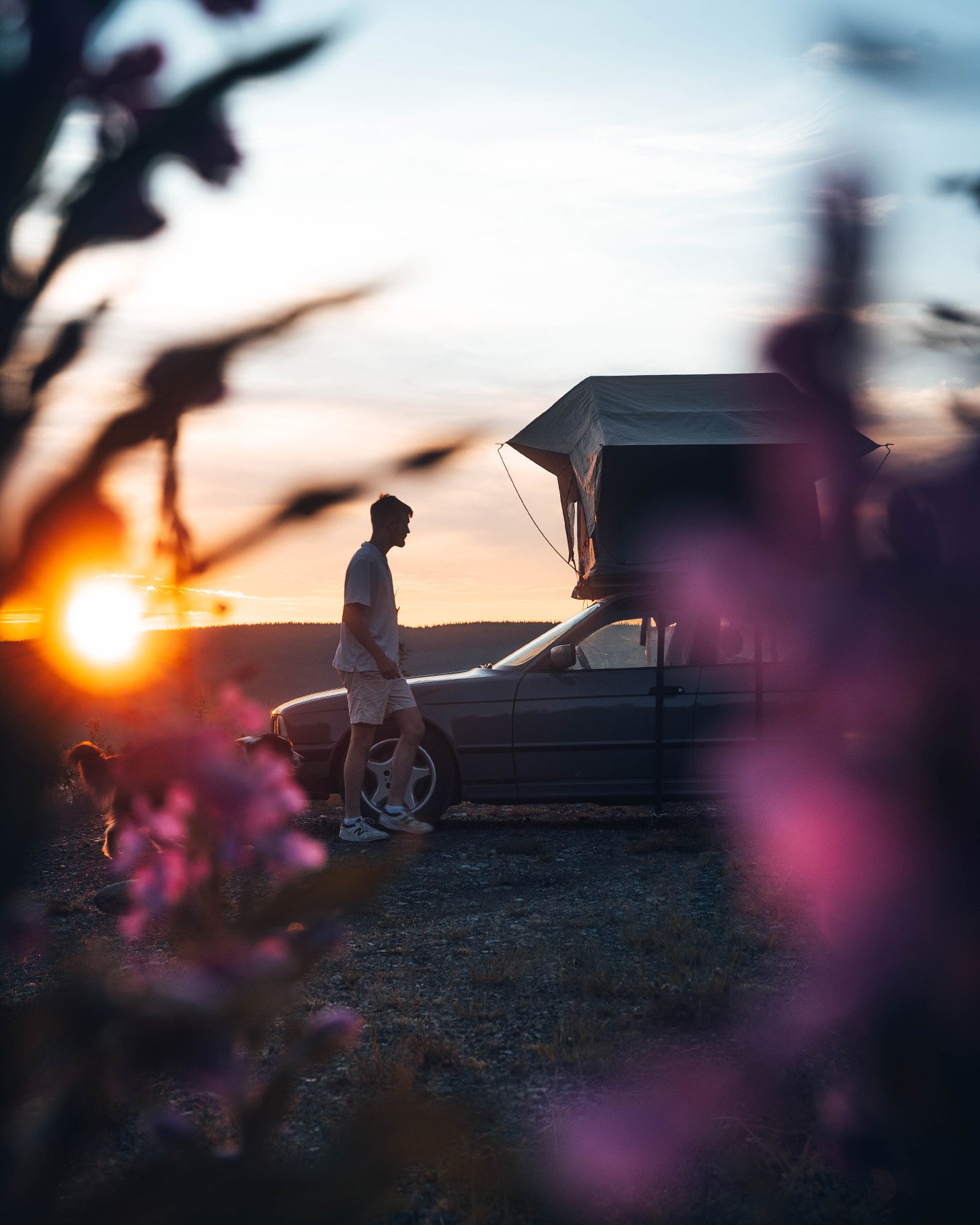 Une tente de toit installée sur un véhicule au coucher du soleil, avec une personne à proximité et des fleurs au premier plan.