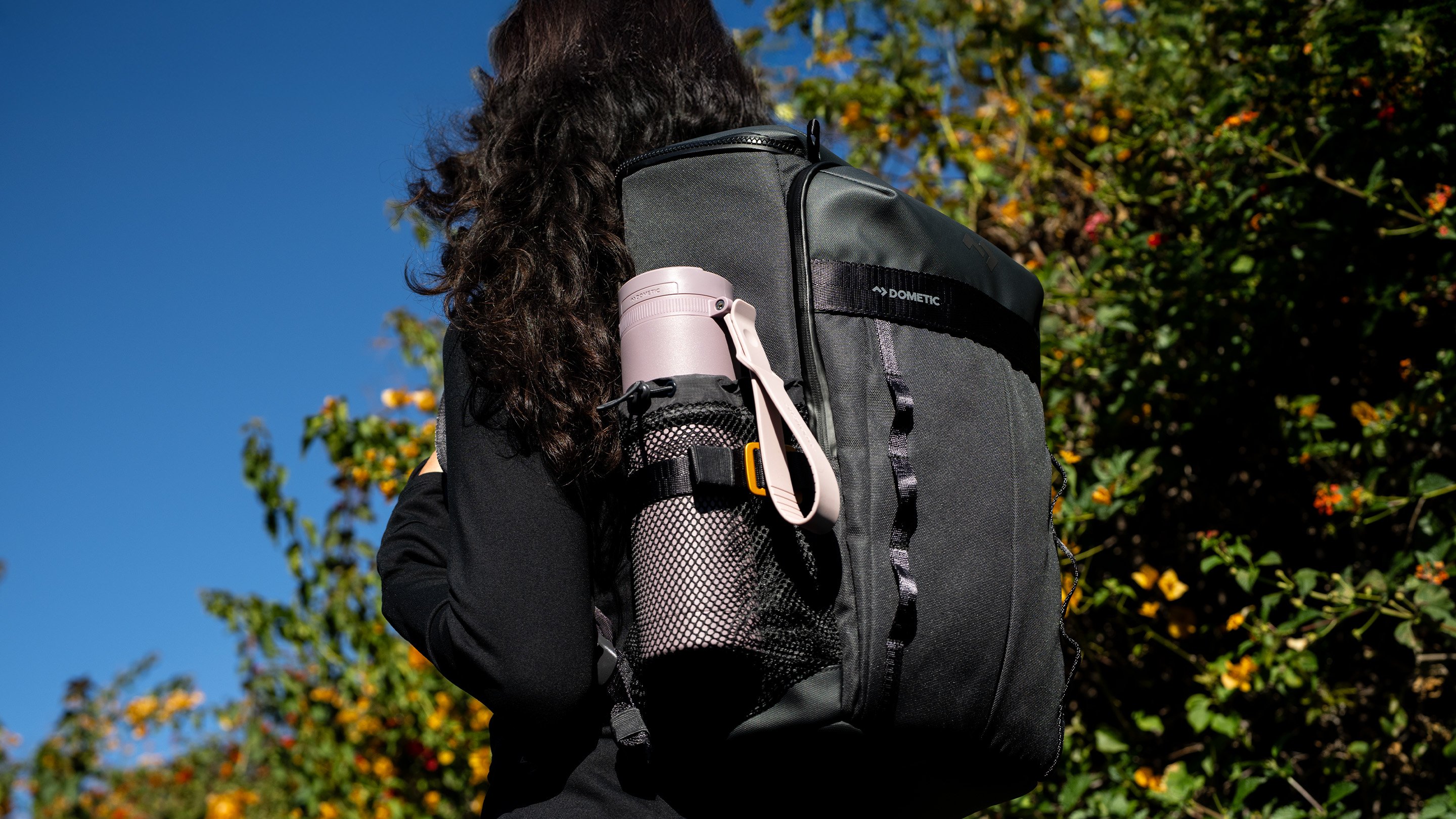 Persona con una mochila negra Dometic al aire libre, con una botella térmica rosa en el bolsillo lateral de malla.
