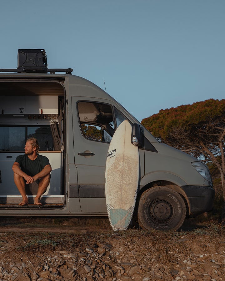 Van camperizzato parcheggiato all’aperto con la porta laterale aperta e una tavola da surf appoggiata al veicolo.
