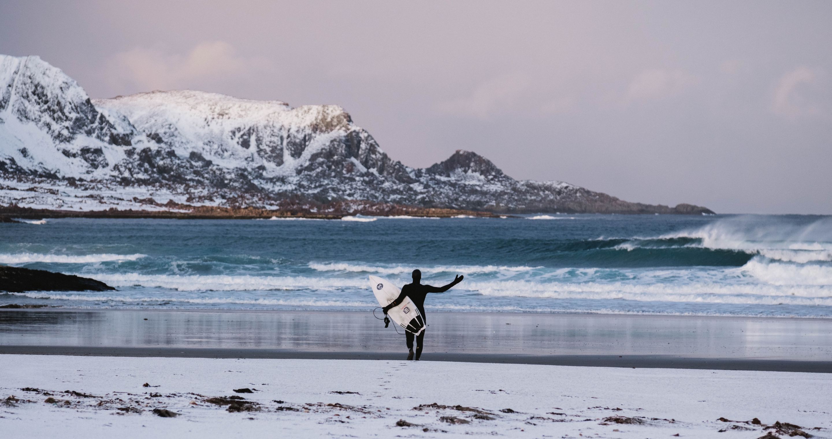 A person in a wetsuit with a surfboard walking towards the ocean with snowy mountains in the distance and pink/purple sky