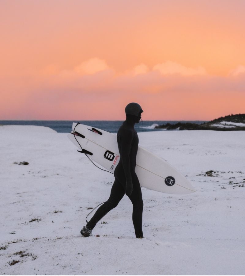 A person in a full wetsuit walking in the snow with a surfboard and sunset skies.