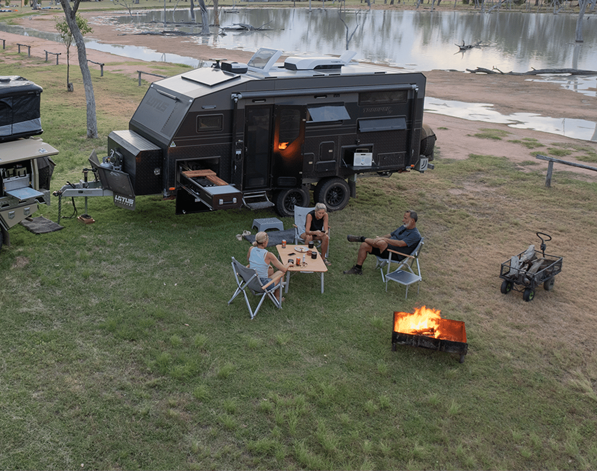 Off-road camper trailer with Dometic RV air conditioner parked beside a lake at dusk, with a campfire and folding chairs.