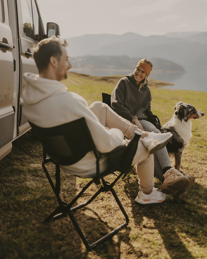 Two people sitting in outdoor chairs beside a camper van with a dog, overlooking a natural landscape.