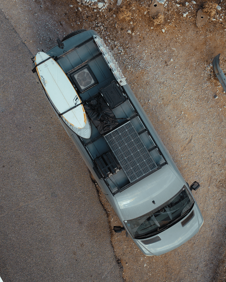 Aerial top‑down view of a vehicle with a surfboard, solar panel, and gear mounted on the roof near a rocky surface.