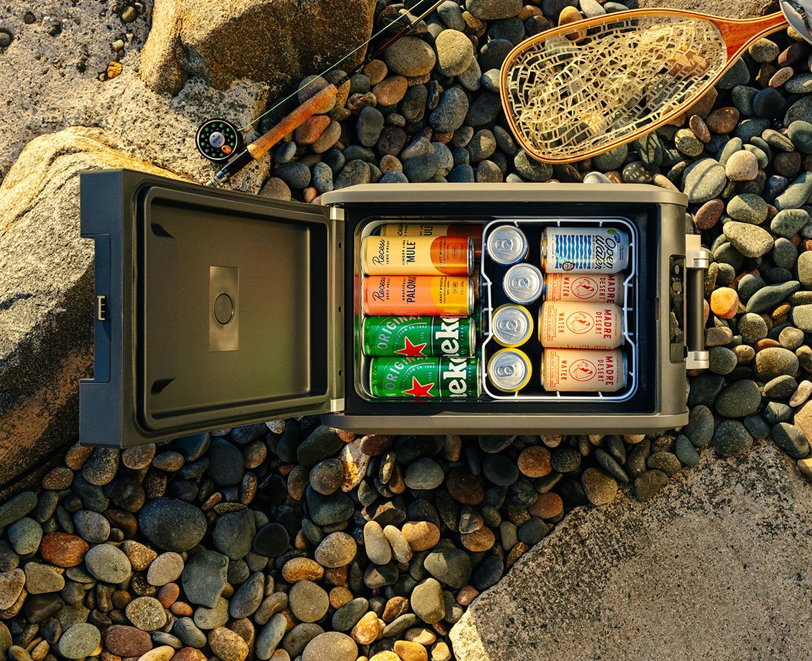 Open Dometic CFX5 compressor cooler filled with beverage cans, portable electric cooler placed on rocky ground outdoors
