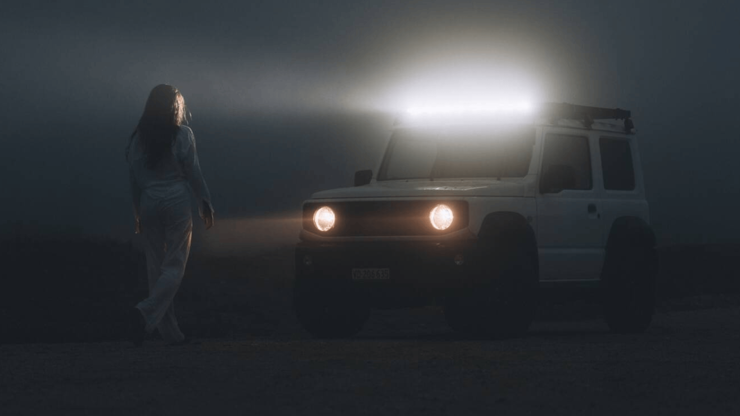 Woman in darkness standing in headlights of white Jimny.