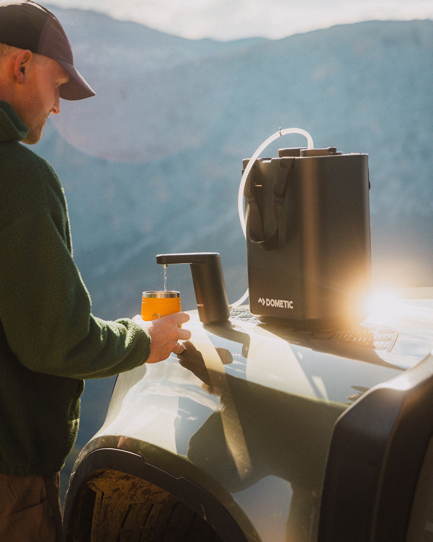 Dometic draagbare watercontainer met kraan die water in een beker tapt op de motorkap van een voertuig in een bergachtige buitenomgeving.