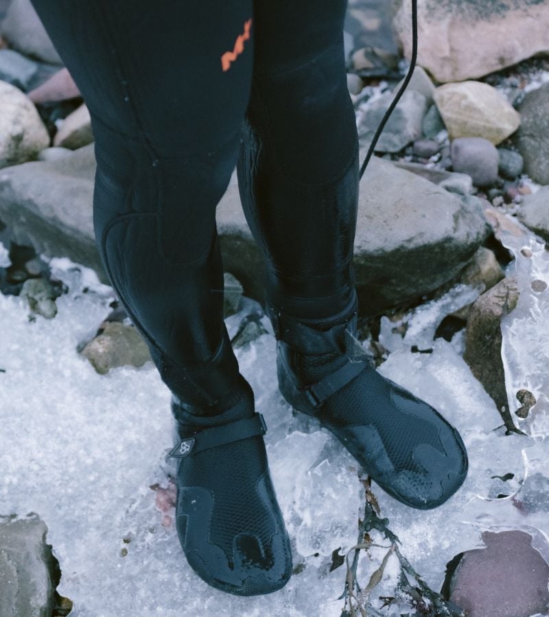 A close up of a person's legs in a wetsuit, standing on ice/rocks.