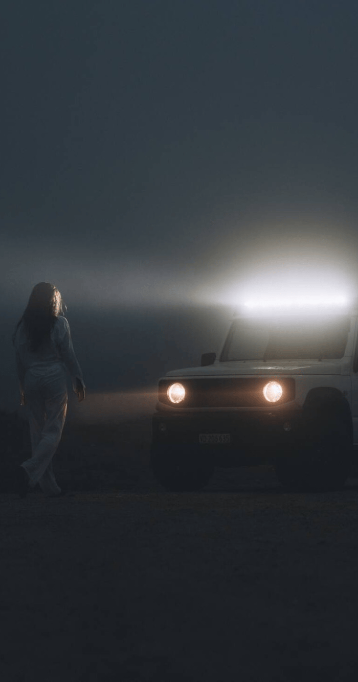 Woman in darkness standing in headlights of white Jimny.