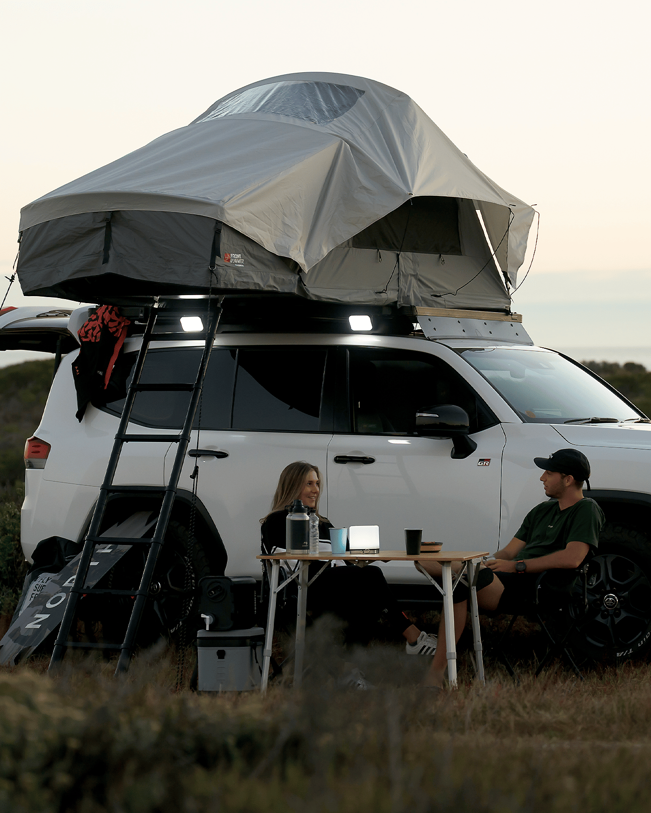 White vehicle near the coast at sunset with a Front Runner Dometic Feather‑Lite II rooftop tent set up, ladder attached, and two people seated at a camping table beside the vehicle.