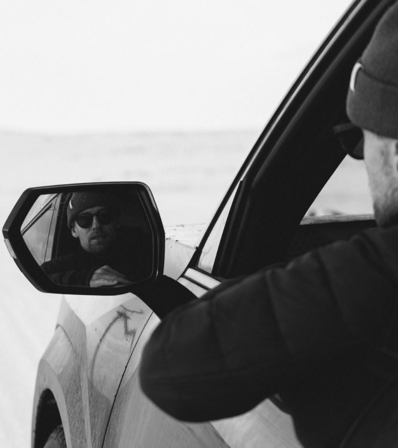 A male leaning out of the driver’s side window of a vehicle, with their reflection visible in the side mirror