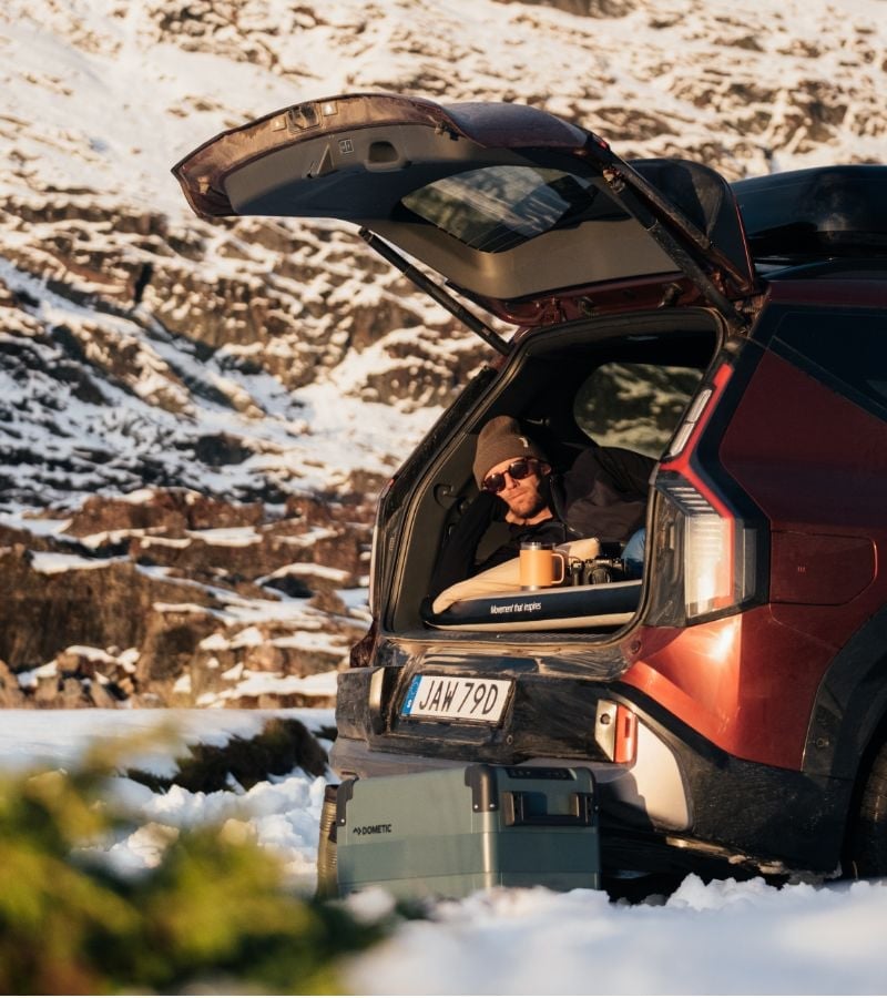 A person relaxing in the back of a SUV with icy mountains in the background and Dometic cooler on the icy ground.