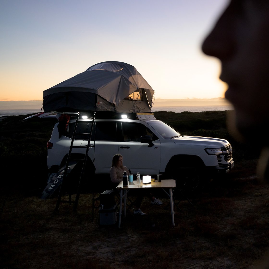 Veicolo bianco parcheggiato sulla costa al tramonto con tenda da tetto Front Runner Dometic Feather‑Lite II montata, scala fissata e tavolo da campeggio accanto al veicolo.