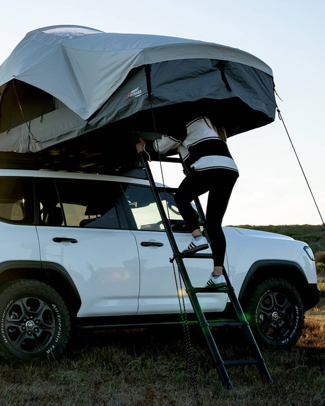 Person climbing the ladder into an opened Front Runner Dometic Feather‑Lite II rooftop tent mounted on a white vehicle outdoors.
