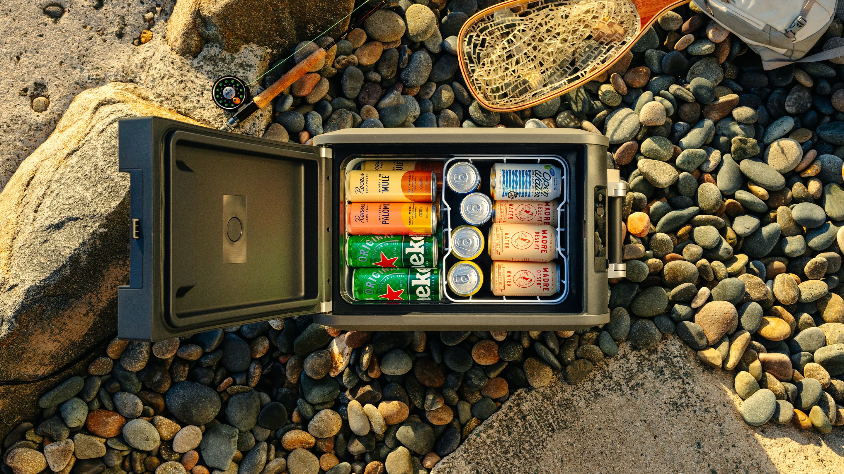 Top‑down view of an open Dometic CFX5 compressor cooler filled with canned drinks, placed on a rocky beach with fishing gear nearby.