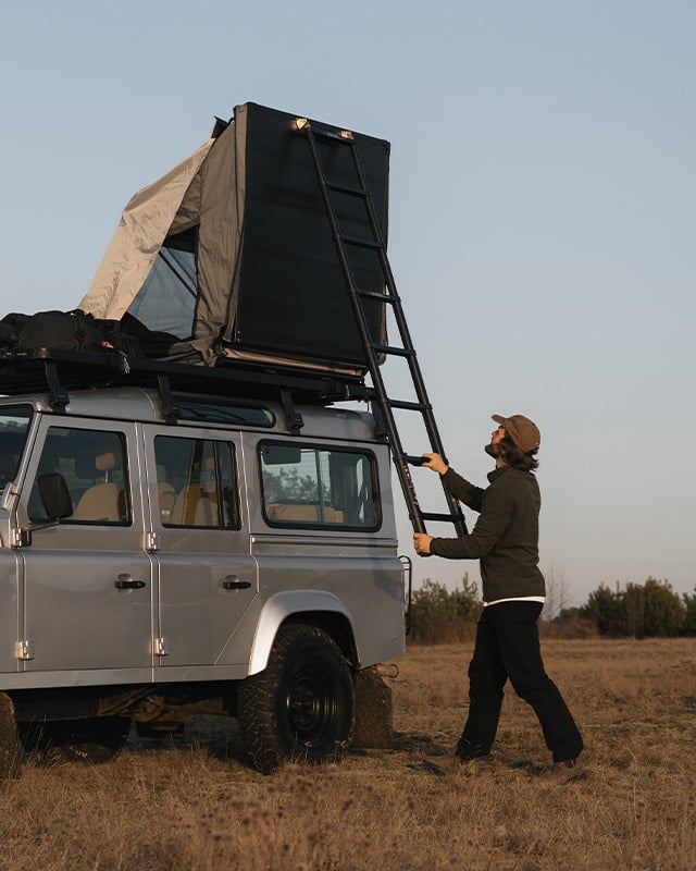 Person closing the Front Runner Dometic Feather‑Lite II rooftop tent using the telescopic ladder on a silver vehicle in an open field.
