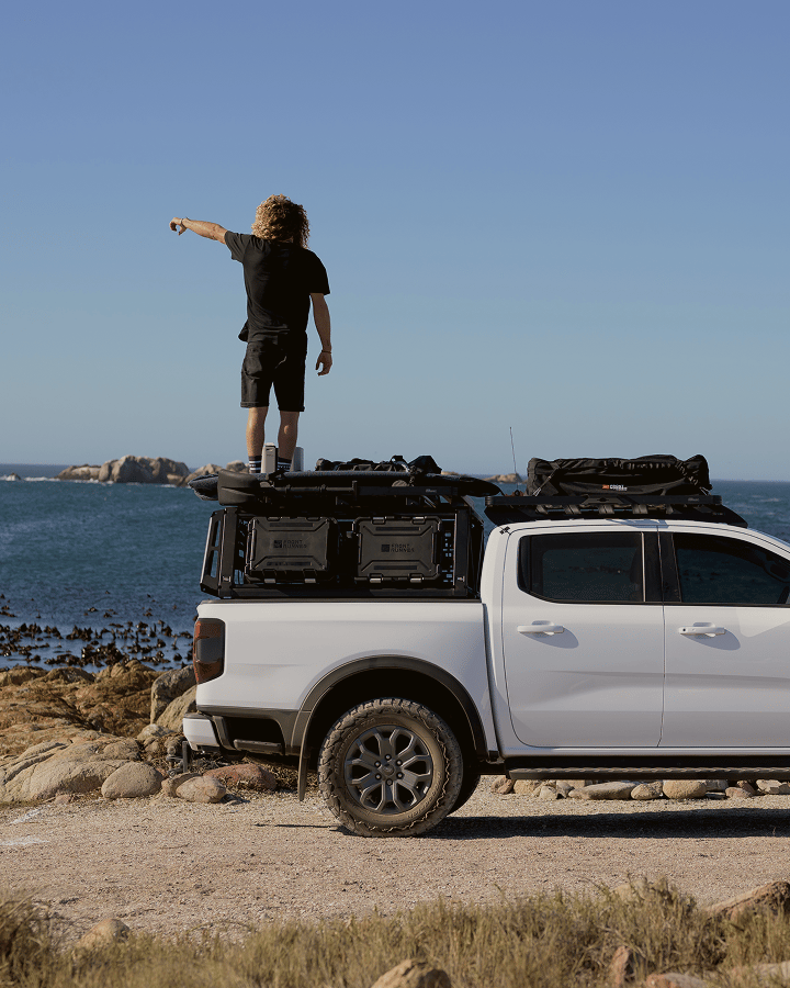 Pickup truck equipped with a Pro Bed Rack carrying storage cases, with a person standing on the rack platform overlooking a coastal landscape.