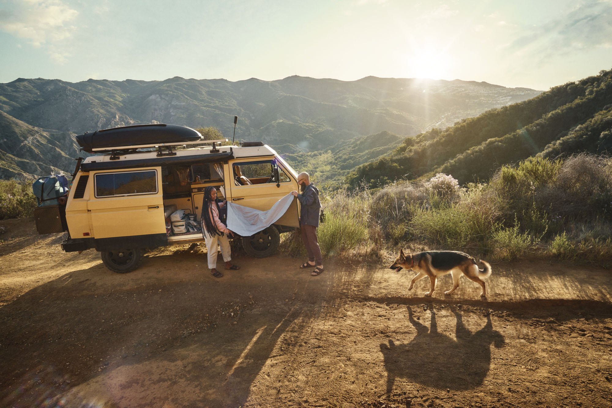 A man and woman folding a blanket outside their old school yellow VW camper van, with a german sherpard dog running near by.