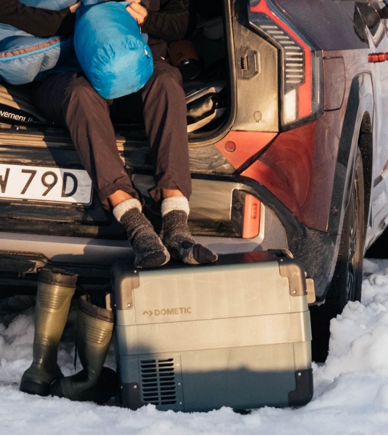 A person sitting in the back of a SUV with their legs resting on a Dometic cooler sitting in the snow. 