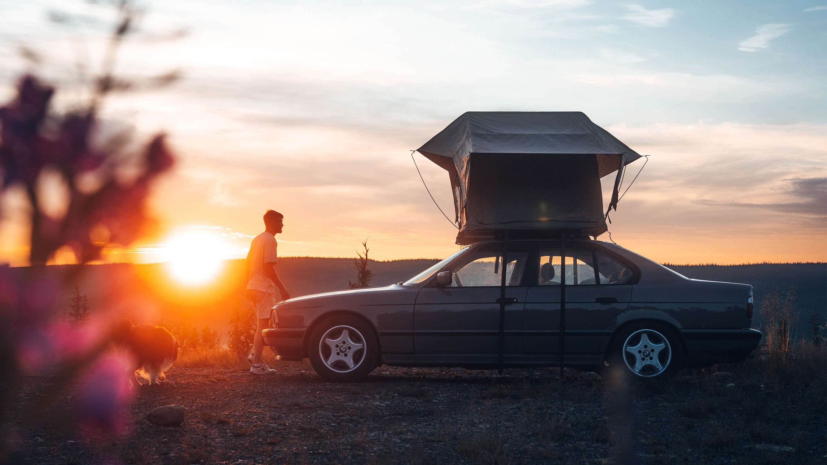 Car with a roof tent set up on rocky terrain at sunset, with a person standing next to the vehicle and soft‑focus flowers in the foreground.