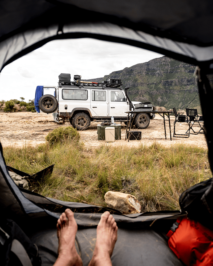 Vue depuis l’intérieur d’une tente sur un véhicule tout‑terrain équipé d’une galerie de toit et d’un campement en plein air.