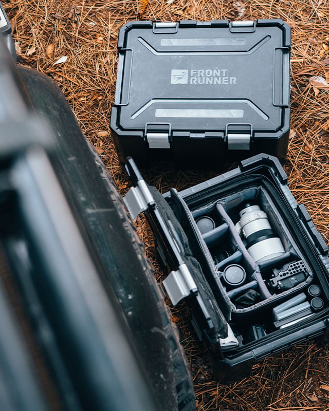 Front Runner Wolf Pack Pro storage boxes on the ground beside a vehicle, with one storage box open to reveal organized camera equipment and accessories