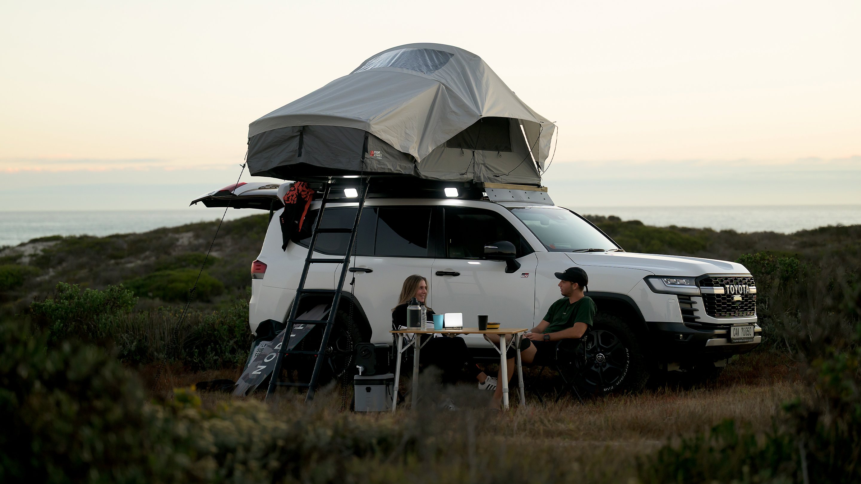 Véhicule blanc près de la côte au coucher du soleil avec une tente de toit Front Runner Dometic Feather‑Lite II installée, échelle fixée et table de camping à côté du véhicule.