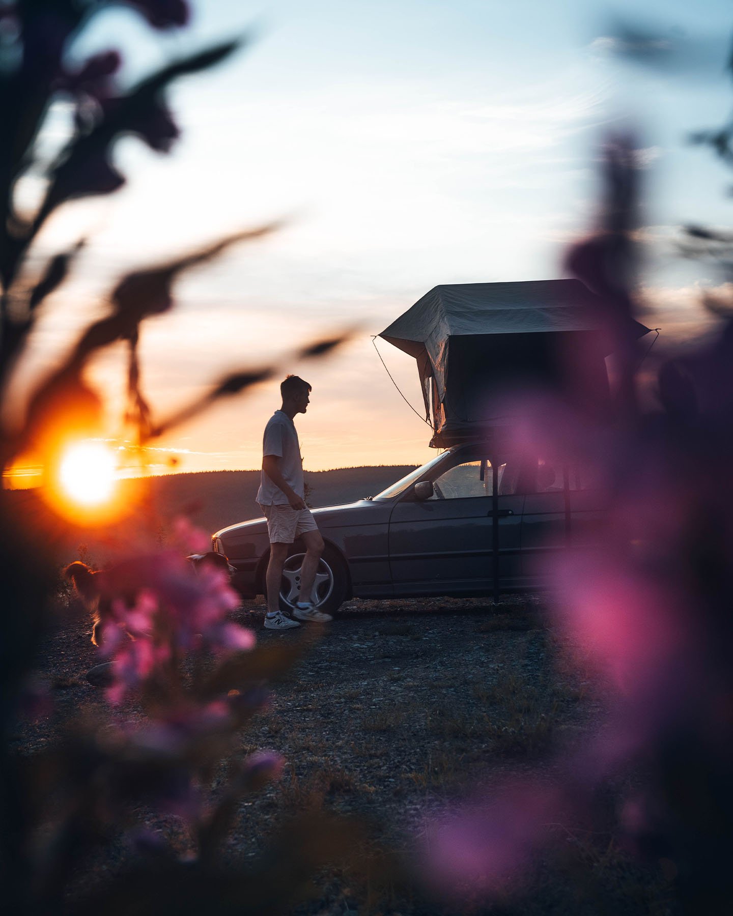 Voertuig met een daktent geparkeerd bij zonsondergang, omlijst door onscherpe bloemen op de voorgrond met een persoon naast de auto.