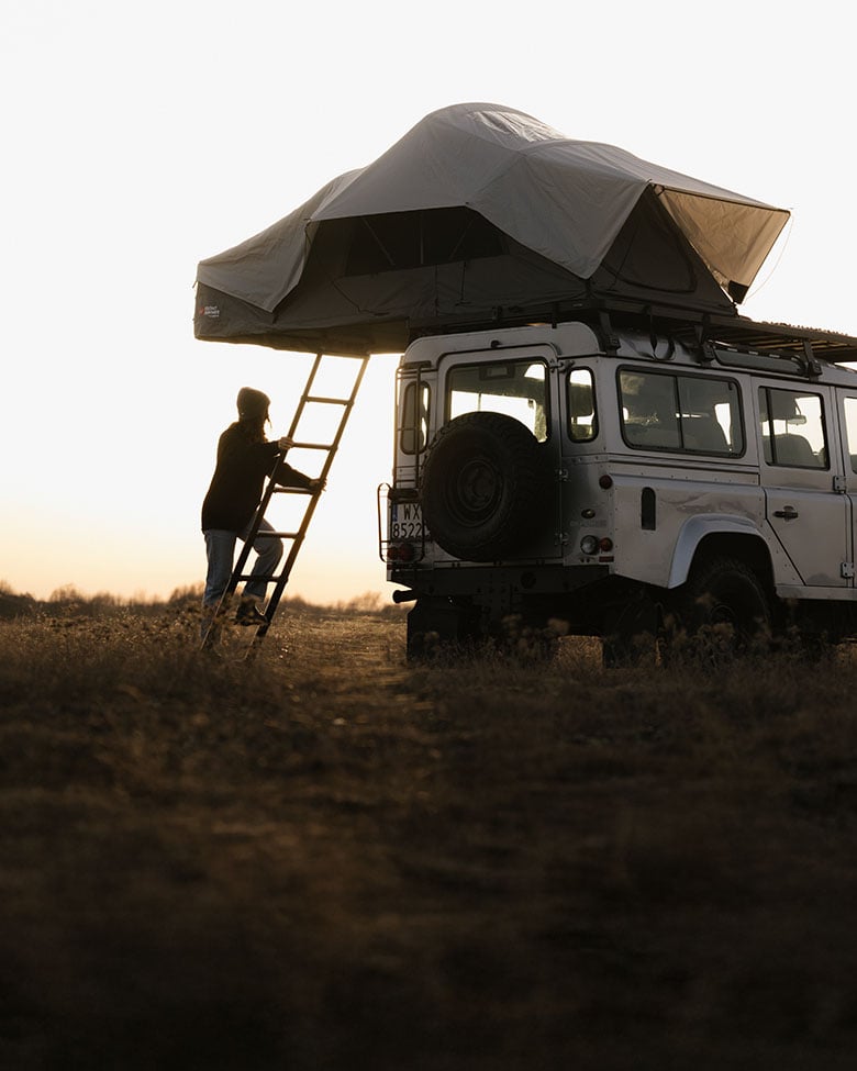 Véhicule tout-terrain avec tente de toit dans un paysage ouvert au coucher du soleil, personne montant à une échelle arrière pour accéder au camping surélevé