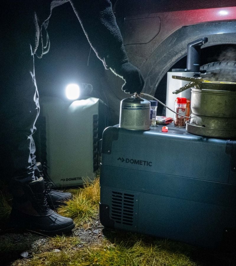 A person cooking at night using a Dometic cooler as a table. A camp light casting a glow on the scene near the back of a vehicle.