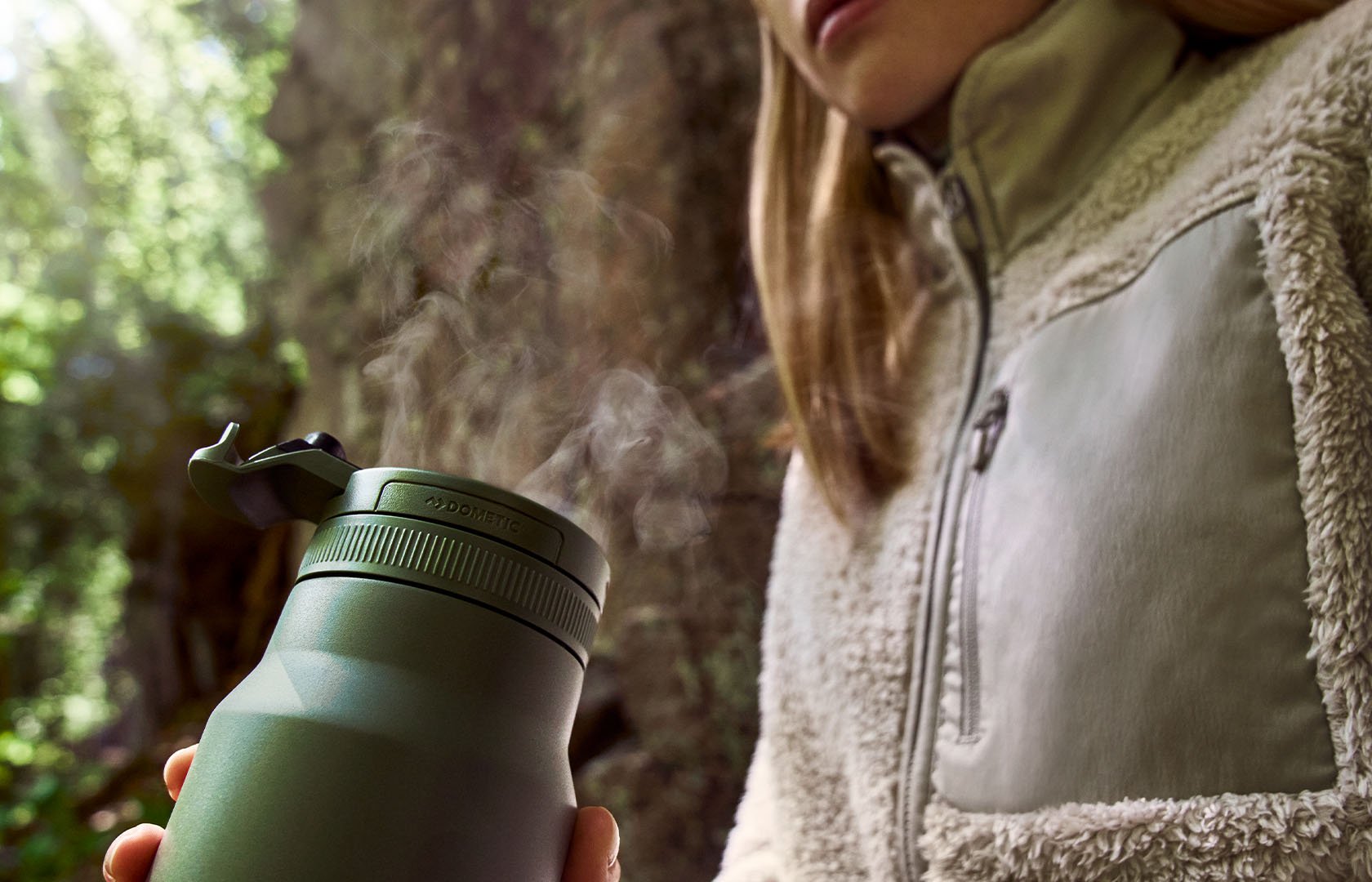 Grön isolerad termoflaska med ånga från locket, hållen utomhus intill ett träd.